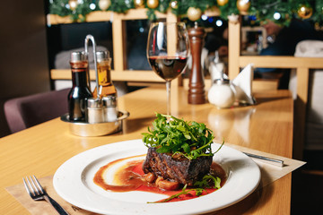 Beef steak with arugula and champignons on a white plate on the table in the restaurant. A glass of red wine, a salt, a pepper.