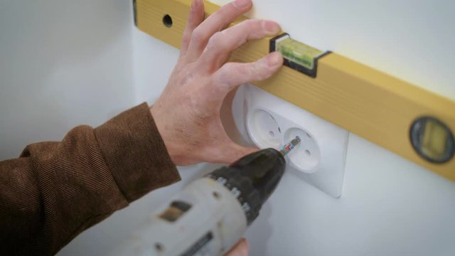 wireman is mounting a double socket on a white wall inside a flat, aligning and twisting bolts by screwdriver
