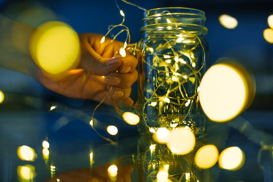 Female hands holding glass mug with garlands of lights .