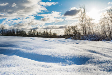 snowy field and forest during the sunset of the bright sun, blue sky with clouds, winter landscape