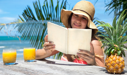 beautiful girl in a straw hat reading a book on a background of palm trees and summer drink with pineapple, on the beach