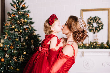 Young beautiful mother holding her infant daughter both wearing red dresses, kissing and hugging by the Christmas tree