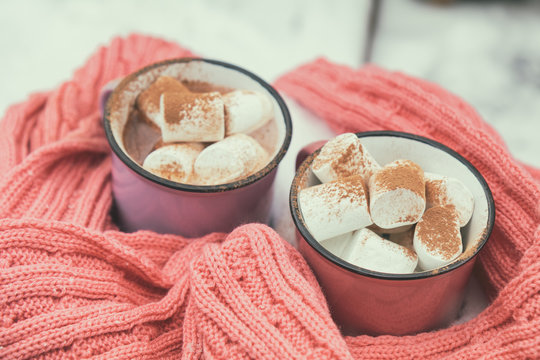 Hot chocolate with marshmallow in pink and violet two cups wrapped in a cozy winter pink scarf on the snow-covered table in the garden. Coloring and processing photo small depth of field