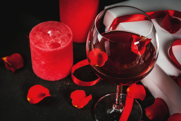 Valentine's day concept, white wrapped gift box with red ribbon, rose flower petals, red wine glass, with red candle, on dark stone background, copy space