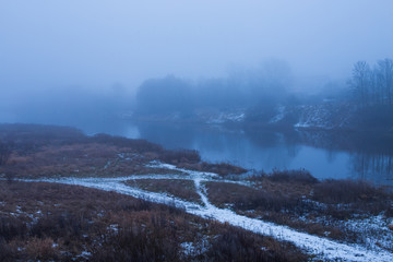 Misty winter evening over the river.  Naked trees in river bank. Snowy roud along the river.