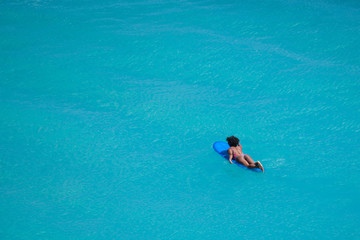 Surfer girl with afro hairstyle paddling out on surfboard in crystal clear turquoise water at Uluwatu beach, Bali, Indonesia