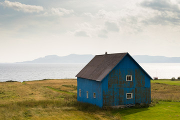Fototapeta na wymiar A blue wooden farmhouse with white window frames standing alone in a field in front of a bay of water along the coastline.