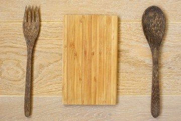 A wooden cutting board and a set of salad utensils laid out on light wooden background. 