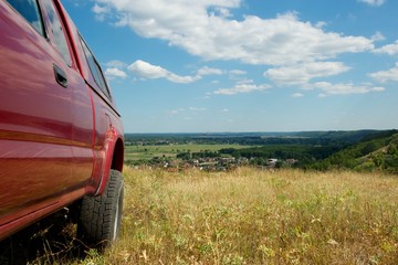 Picture taken from behind a rear wheel of a red pickup truck. Beautiful view from a mountain top. Dry grass, cloudy sky. © Oleksii Klonkin