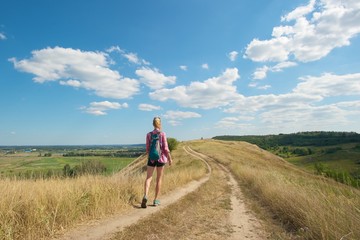 A young woman walking on dry grass up a hill her back turned to the camera; wearing short shorts, plaid shirt and matching color trekking shoes and backpack. 