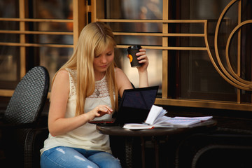 Focused blonde woman copywriter sitting on cafe terrace with coffee. Woman freelancer typing on a...