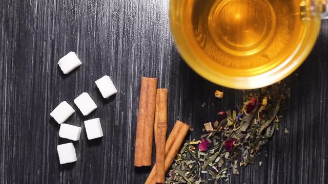 Glass cup of tea with sticks of cinnamon, dried leaves of tea and sugar cubes on the dark wooden surface.
