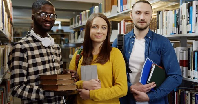 Portrait of three young multi ethnical students standing in front of the camera with books and tablet device in their hands in the library. Indoors