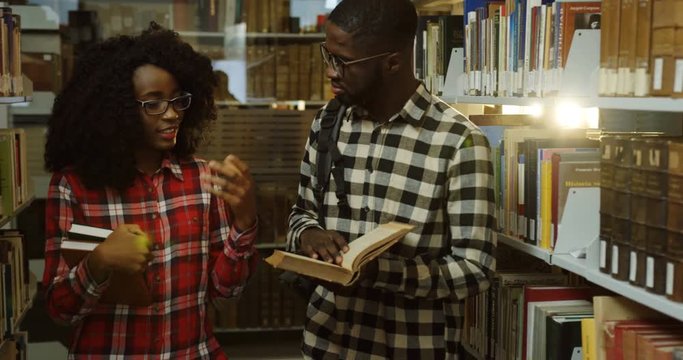 African American couple of students in motley shirts walking through library passages with books in hands and talking. Indoors