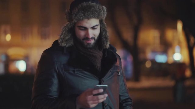 Handsome young man in a stylish winter outfit stands outside and uses his cell phone for surfing the internet or texting messages, smiles towards the screen. Modern trends, contemporary culture