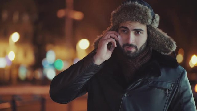 Handsome bearded man in a stylish leather jacket and fur hat walking down the winter street, dials the number and talks on his phone. Winter season, cold weather. Successful lifestyle, active life