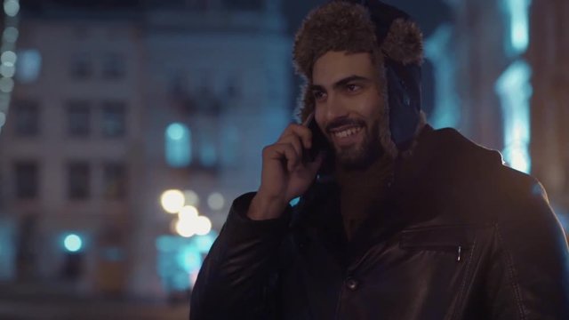 Close up view of young bearded man in a warm leather jacket and fur hat cheerfully communicating via his smartphone. Positive emotions, being happy, holiday atmosphere