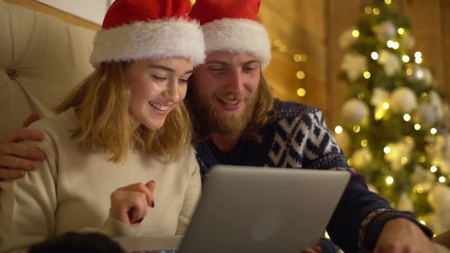 Portrait of young man and woman in Santa Claus red hats skyping greeting friends using laptop while resting in cozy room with Christmas decorated tree
