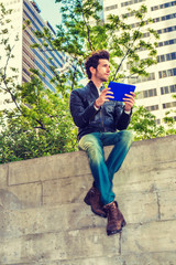 Power of Reading.  A young guy reading small, blue tablet computer on the top of wall on business district. Green trees, strong sun shine on background. Technology in our daily life. Filtered effect..