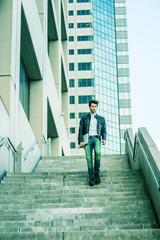 Young man with beard, wearing leather jacket, jeans, arm carrying laptop computer, walking down stairs outside office high building after busy working day, going home. Filtered look with green tint..