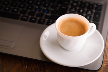 Cup of coffee on a laptop keyboard. Work place modern. Laptop (notebook) with cup of coffee and notepad with pen on old wooden table.