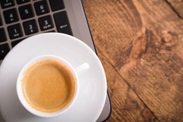 Cup of coffee on a laptop keyboard. Work place modern. Laptop (notebook) with cup of coffee and notepad with pen on old wooden table.