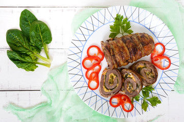 Delicious meat roll with spinach, cheese and pickled cucumber on a plate on a white wooden background.