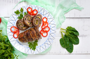 Delicious meat roll with spinach, cheese and pickled cucumber on a plate on a white wooden background.