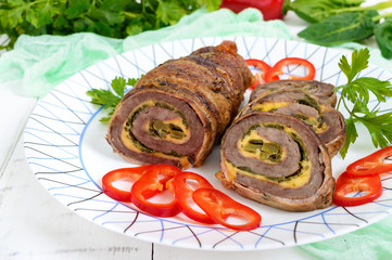 Delicious meat roll with spinach, cheese and pickled cucumber on a plate on a white wooden background.