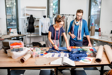 Couple of fashion designers working with fabric and clothing sketches at the studio full of...
