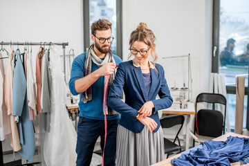 Handsome tailor measuring jacket on the woman client standing at the studio full of tailoring tools and equipment - Powered by Adobe
