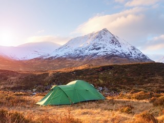Green touristic tent on meadow at river below snowy cone of mountain Stob Dearg 1021 metres high