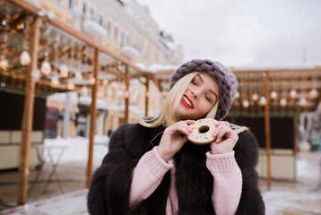 Amazing blonde woman dressed in fur coat  and knitted hat, holding tasty christmas gingerbread...