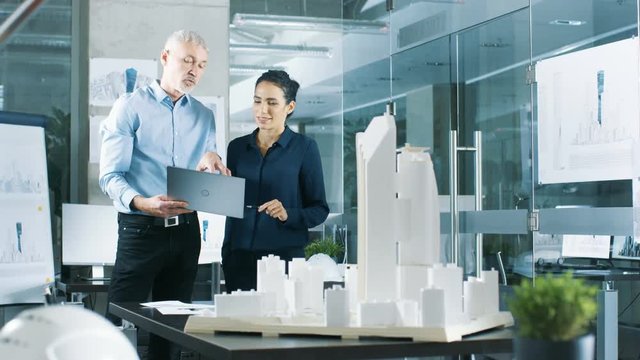 Male Senior Engineer Holds Laptop and has Discussion with Female Chief Engineer. They Work on a Building Model for a New District Urban Planning Project. Shot on RED EPIC-W 8K Helium Cinema Camera.