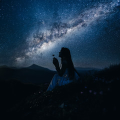 Woman looking at beautiful milky way. Beautiful woman in a long white dress in the mountains. Girl sitting on a rock. Milky Way at mountains. Night colorful landscape. Starry sky with hills at summer