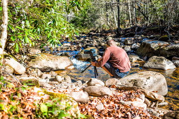 Young man nature photographer with camera and tripod setting up by shallow rock stream with running water and waterfall in autumn with foliage, stones and flowing creek