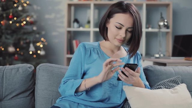Cute woman uses smartphone sitting on the sofa in home smiling portrait close up apartment christmas communication christmas tree hold house internet modern technology rest message slow motion