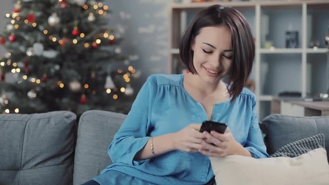 Young woman uses smartphone sitting on the sofa in home smiling portrait close up apartment christmas communication christmas tree hold house internet modern technology rest message slow motion