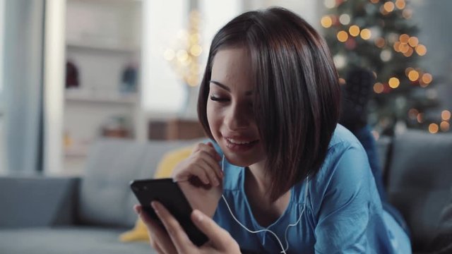 Happy young woman listening to music on phone at home with headphones smiling feels pleasure lying on the sofa attractive relaxation technology apartment modern resting leisure mobile