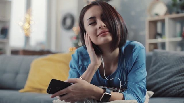 Charming young woman relaxing listening to music on phone at home with headphones smiling feel happy lying on the sofa attractive relaxation technology apartment modern resting leisure mobile