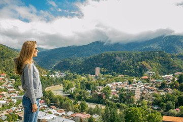 So a beautiful woman is standing on top of a mountain in a hoodie. looking at the mountains and trees from above.Beautiful panorama. Borjomi. Far away city. On top of the mountains and clouds