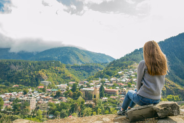 So a beautiful woman is standing on top of a mountain in a hoodie. looking at the mountains and trees from above.Beautiful panorama. Borjomi. Far away city. On top of the mountains and clouds