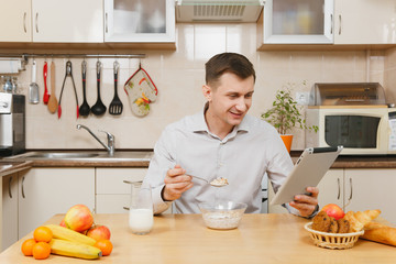 Handsome caucasian young business man in plaid shirt having breakfast, sitting at table with tablet, eating cereals with milk on light kitchen. Healthy lifestyle. Cooking at home. Prepare food.