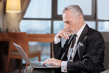 Business in future. Serious senior mediative man looking at the laptop which standing in front of him and thinking 