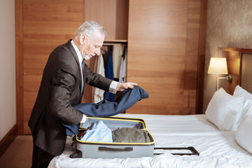 Time to go. Pleasant concentrated senior man standing near the bed placing clothes in the suitcase while staring down