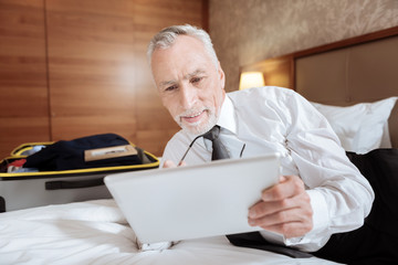 Digital literature. Glad senior merry man relaxing on the bed while carrying tablet and smiling