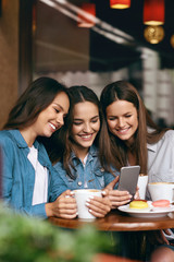 Young Female Friends Using Phone And Drinking Coffee In Cafe.