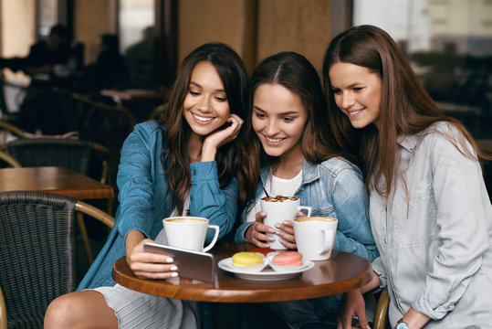 Young Female Friends Using Phone And Drinking Coffee In Cafe.