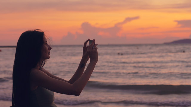 Young tourist woman photographs ocean view with smartphone during sunset at beach