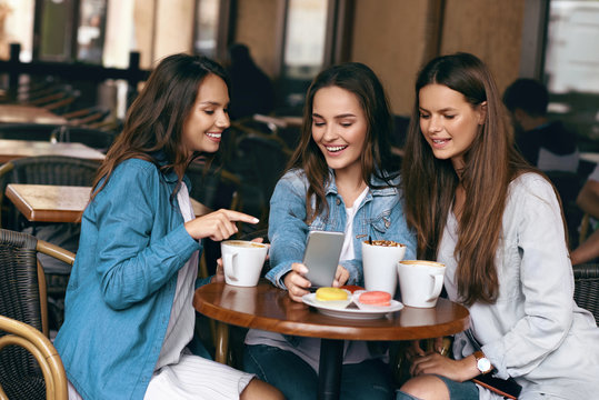 Young Female Friends Using Phone And Drinking Coffee In Cafe.